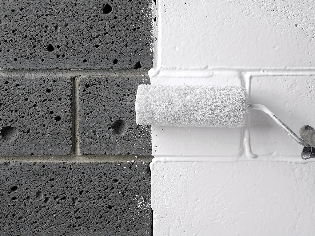 Applying the first coat of acrylic primer to a rough cinder block wall with a roller.