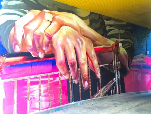 A detailed close-up shot of two pairs of hands, one holding a spray can and the other a detailed stencil, working together on a vibrant graffiti mural.