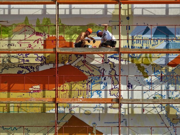 Artists on elevated platforms meticulously preparing a large urban wall for a mural, showcasing cleaning tools, scaffolding, and protective gear.