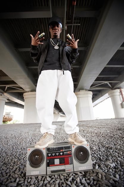 A vintage photograph of a 90s hip-hop artist, possibly wearing an oversized FUBU jersey and baggy jeans, with a gold chain, performing on stage with a microphone.